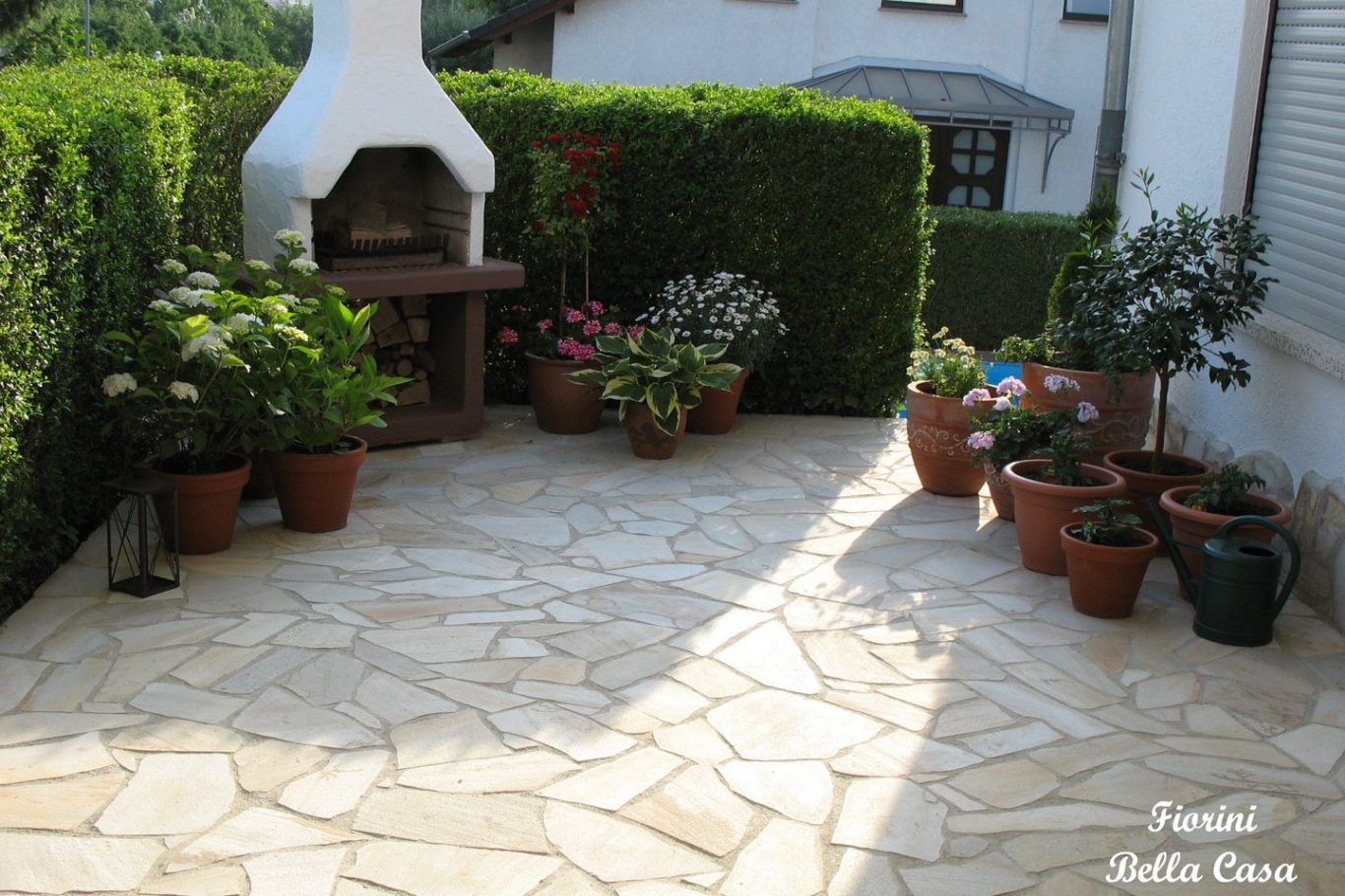 Terrasse mit Porphyr, Blumentöpfen und einem Grill im Freien, umgeben von Hecken.