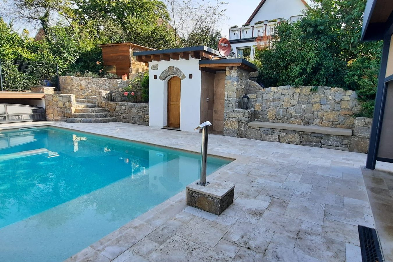 Weißer Pool mit Steinumrandung, umgeben von grünen Bäumen und einer Gartenterrasse.