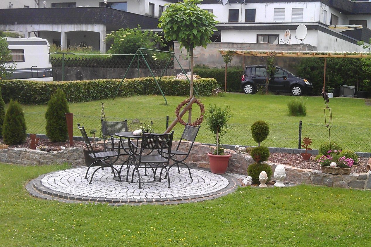 Gemütlicher Garten mit runder Steinterrasse, Tisch und Stühlen, umgeben von Pflanzen.