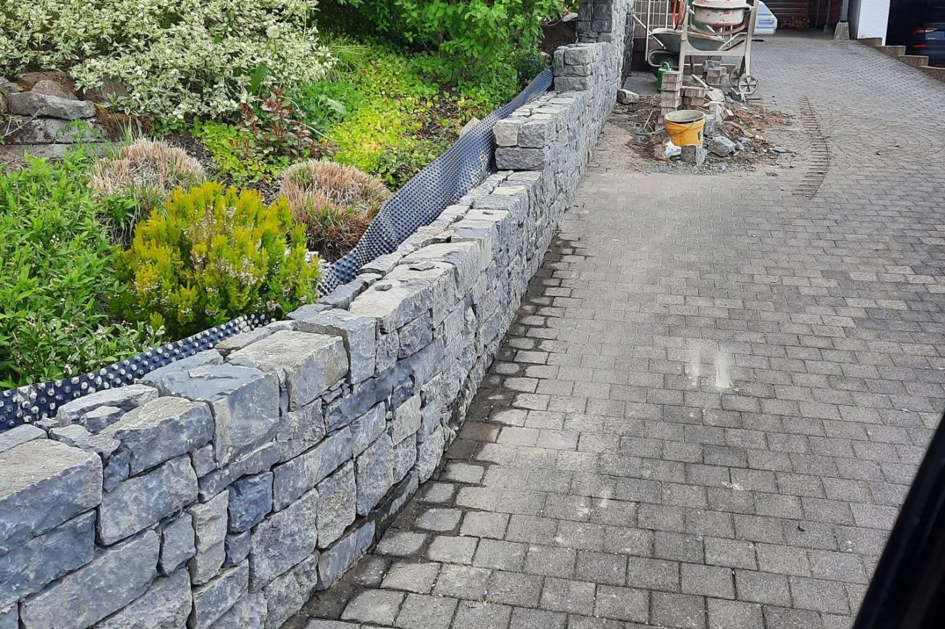 Mauer aus grauen Steinen umschließt einen gepflasterten Weg und Gartenbereich.