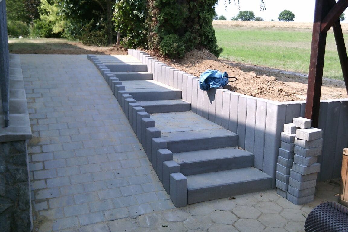 Gartenbereich mit gestuften Betonstufen, Rampenaufgang und Pflastersteinen, umgeben von grünen Wiesen.