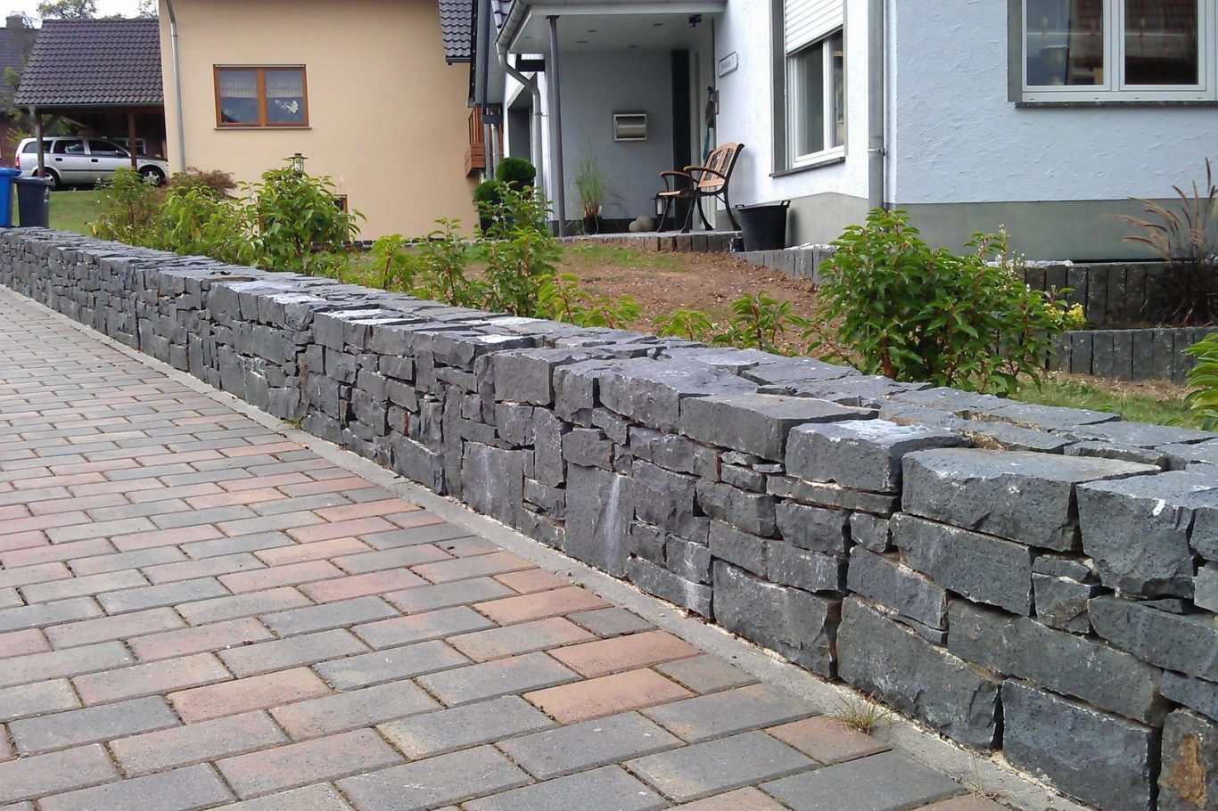 Mauer aus Basaltsteinen entlang eines gepflasterten Wegs mit Betonpflaster vor Wohnhäusern.