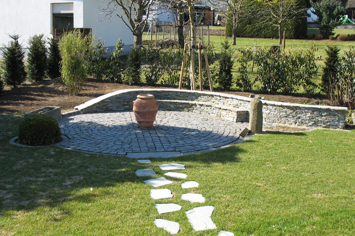Steingarten mit gepflastertem Bereich als Sitzecke, einem Topf und Rasenflächen im Hintergrund.