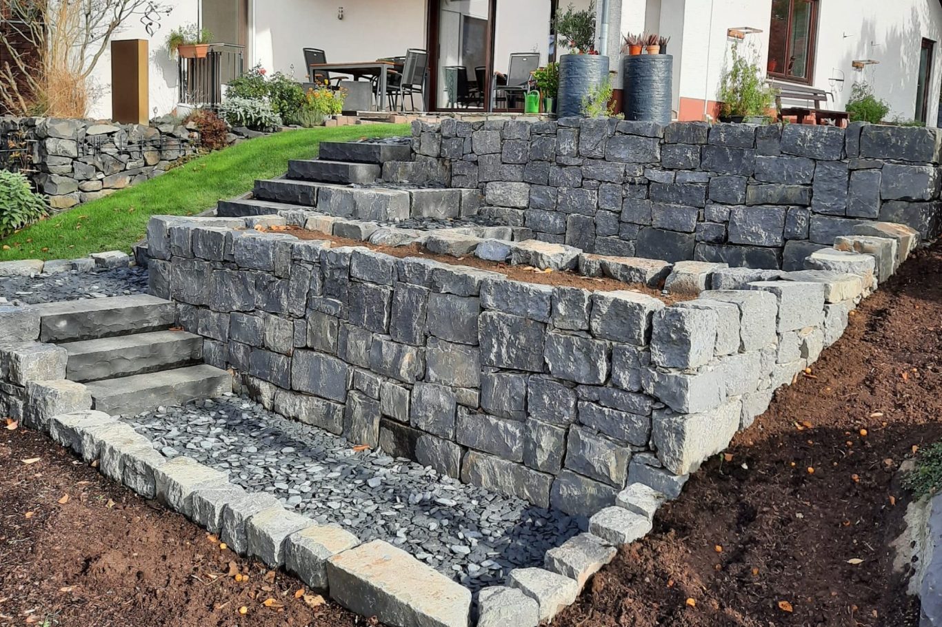 Gestufte Steinmauer am Hang mit Treppen und bepflanzbaren Bereichen für Kräuterbeet im Garten.