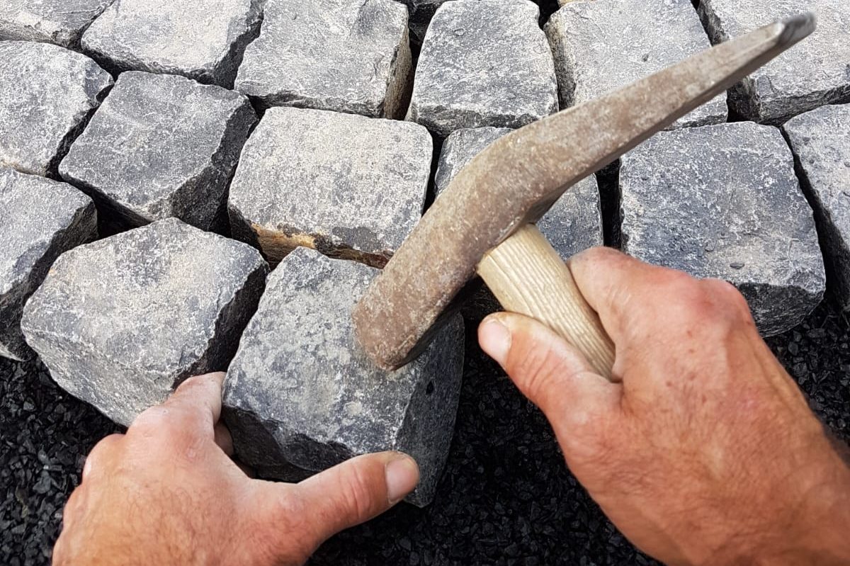 Hände, die mit einem Hammer Steine in einer Pflastersteinstraße bearbeiten.
