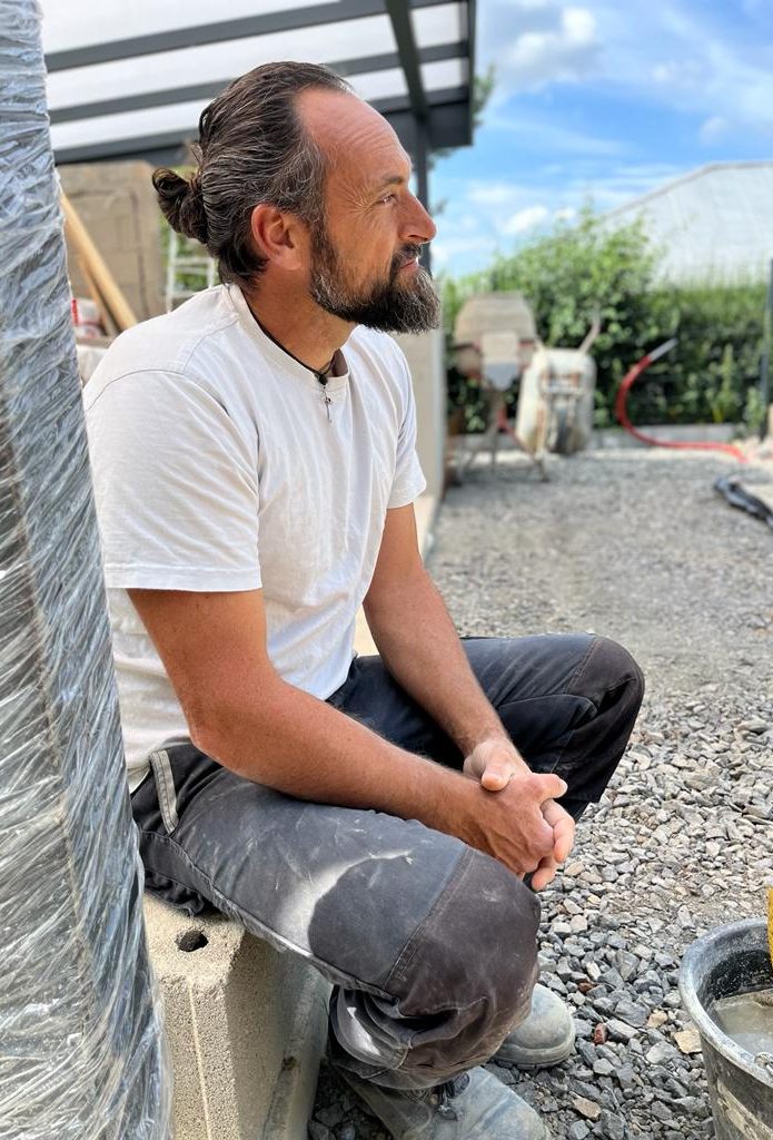 Ein Mann in Arbeitskleidung sitzt außerhalb, umgeben von Baustellenmaterialien.
