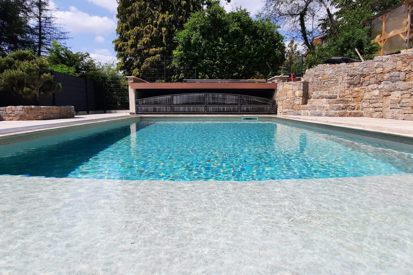 Klarer Pool mit glitzerndem Wasser, umgeben von Natur und Steinmauern.