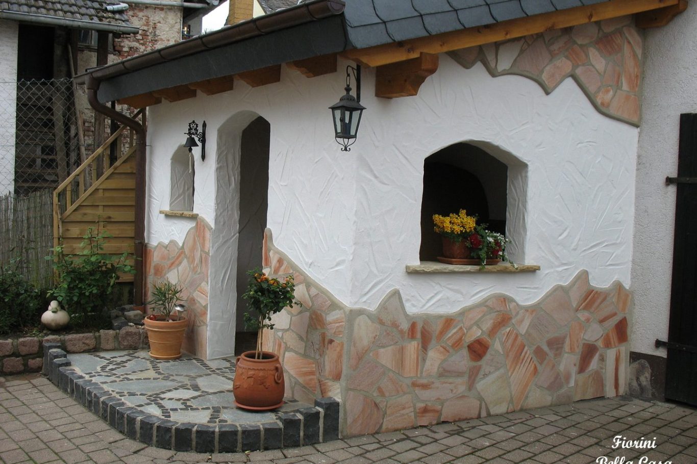 Hauswand mit dekorativem Putz, Fenstern und Blumen auf der Fensterbank. Kleinpflanzen davor.
