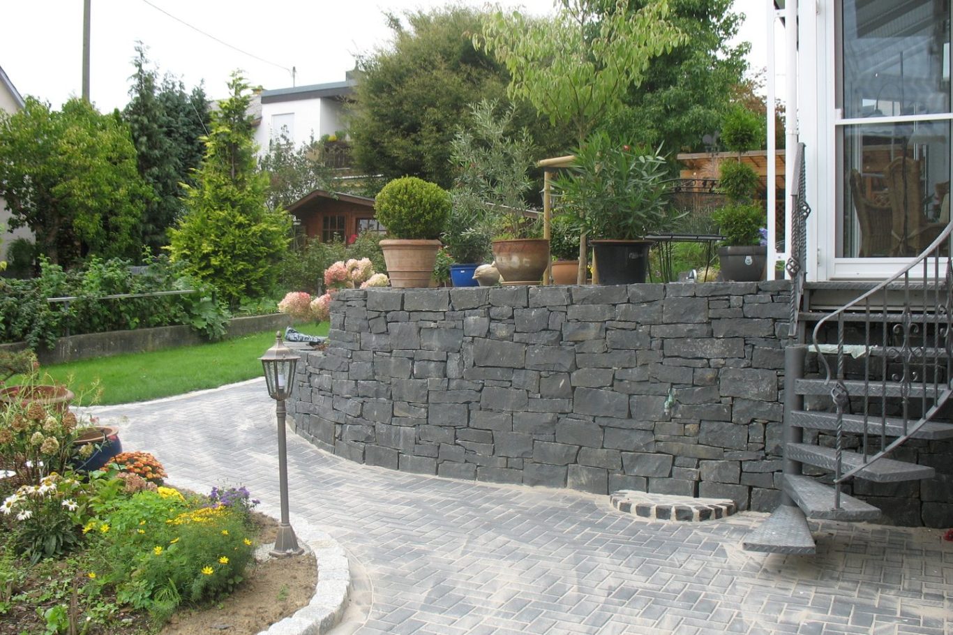 Gartenansicht mit Steinmauer, Treppe und bepflanzten Töpfen.